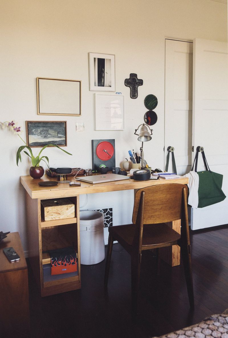 Interior room with a wooden desk, chair, laptop, and decorative items, set against a white wall with framed art.