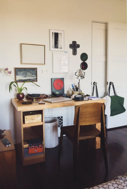 Interior room with a wooden desk, chair, laptop, and decorative items, set against a white wall with framed art.