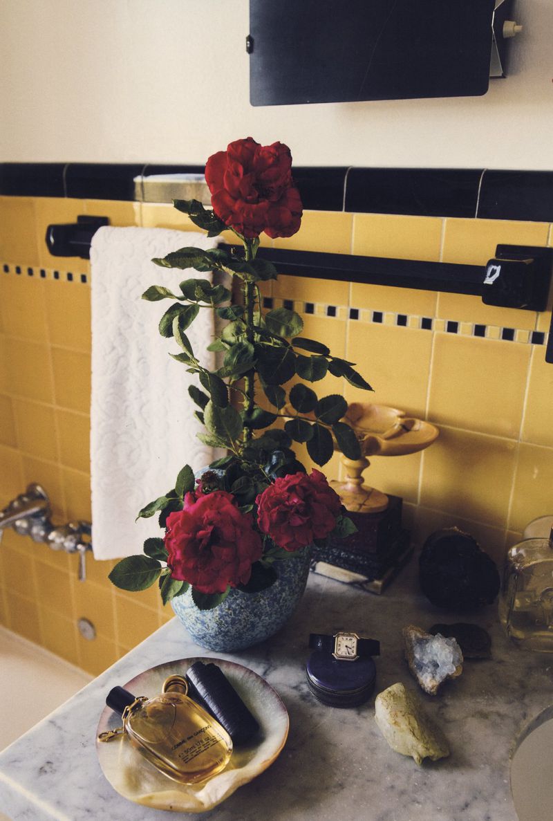 Arrangement with red roses and beauty products on a marble bathroom counter with yellow tile and black accents.