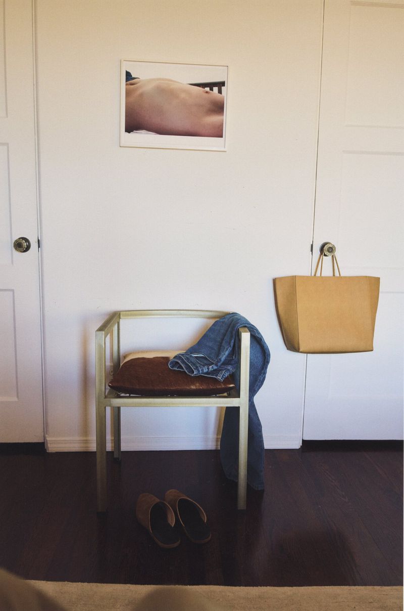 Indoors showing a light gold chair with blue jeans, a brown bag, brown slippers and a framed photo on a white wall.