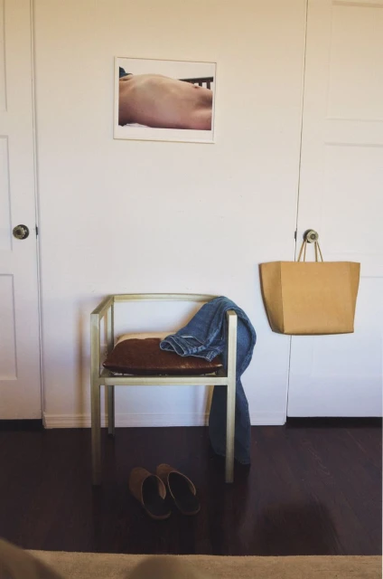 Indoors showing a light gold chair with blue jeans, a brown bag, brown slippers and a framed photo on a white wall.