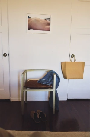 Indoors showing a light gold chair with blue jeans, a brown bag, brown slippers and a framed photo on a white wall.