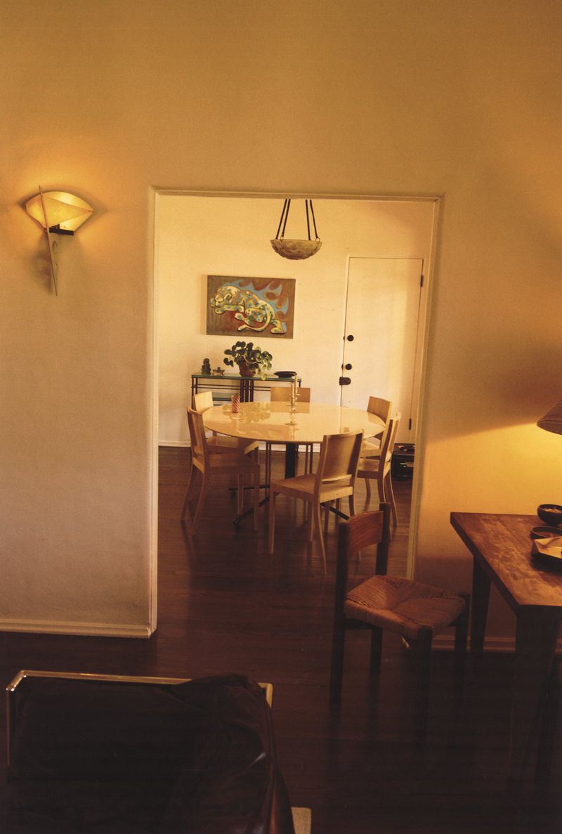 Interior shot of a dining room table and chairs visible through a doorway, featuring a wall-mounted light and dark hardwood floors.