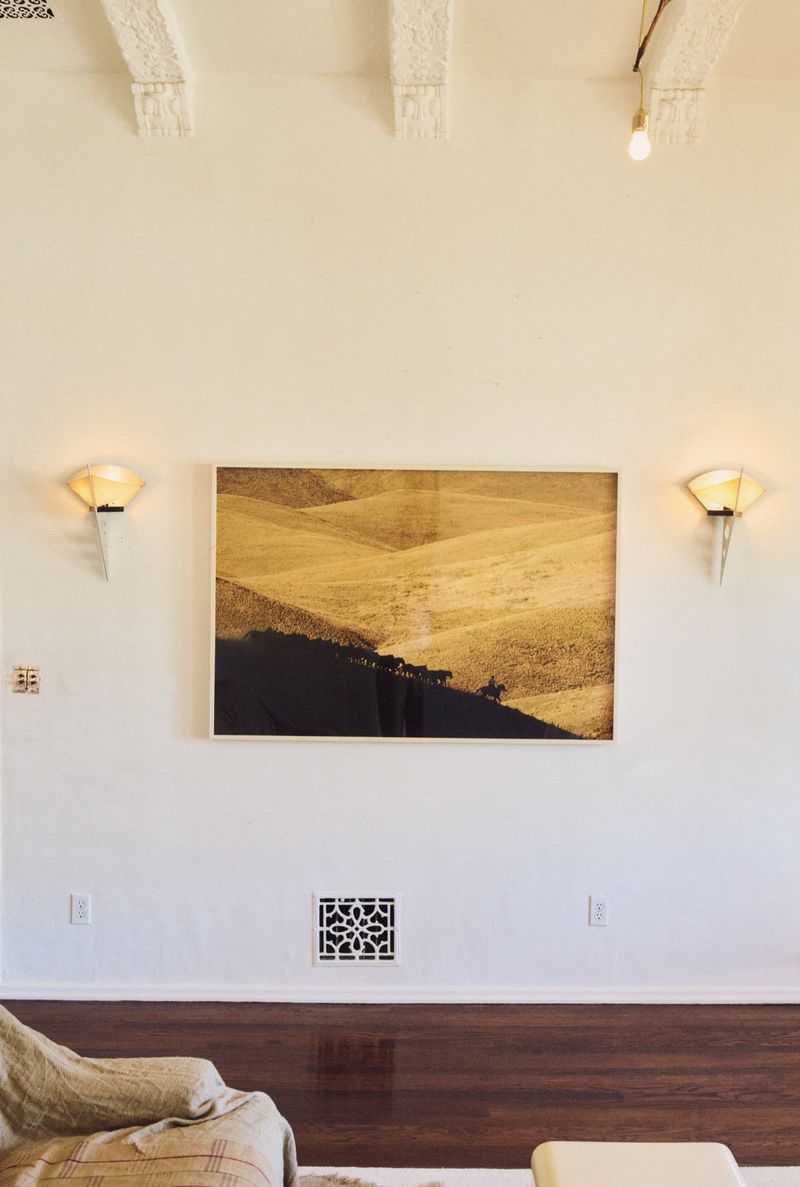 Framed sepia-toned landscape art on a white wall between sconces, above dark wood trim in an interior room setting.