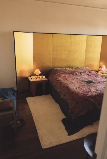Interior bedroom view with gold headboard, patterned bedding, bedside table with lamp, and a neutral-toned rug on a dark wood floor.