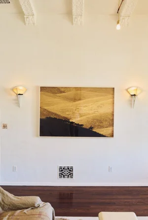 Framed golden landscape art mounted on a white wall flanked by two lighted sconces above dark hardwood flooring.
