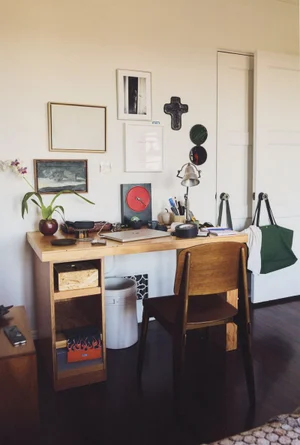 Wooden work desk with a laptop, lamp, and chair placed against a white wall adorned with framed art and hanging items.