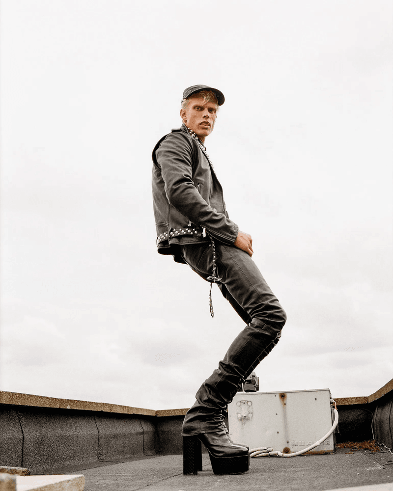 A slender adult male in black leather jacket and platform boots strikes a fashion pose on a rooftop against an overcast sky.
