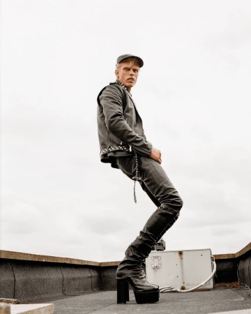 A slender adult male in black leather jacket and platform boots strikes a fashion pose on a rooftop against an overcast sky.