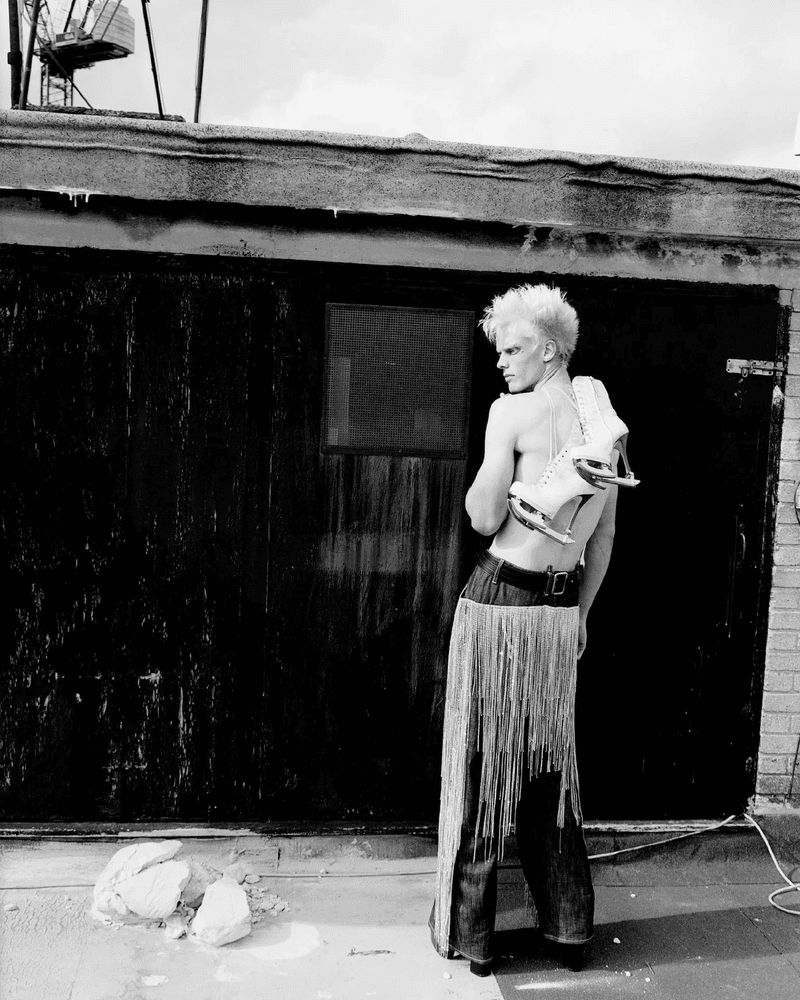 Black and white portrait of a shirtless person with spiky blonde hair and ice skates, posing outdoors in front of a dark building.