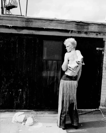 Black and white portrait of a shirtless person with spiky blonde hair and ice skates, posing outdoors in front of a dark building.