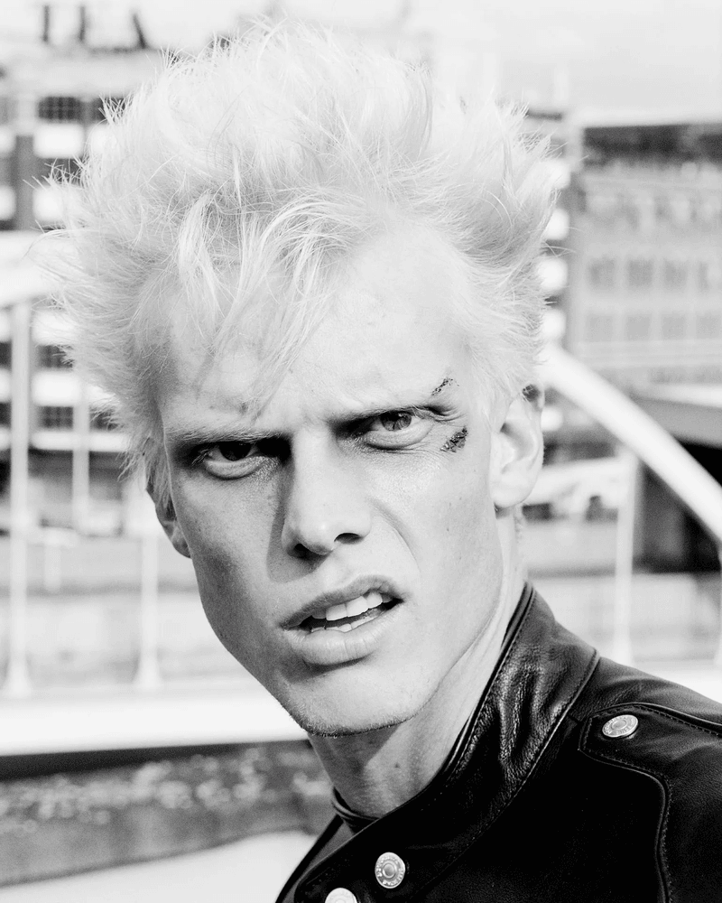 Monochrome portrait of a male model with platinum blonde hair, a leather jacket, and a determined expression in an urban setting.