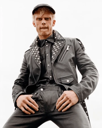 Low-angle shot of a blond adult male wearing a black studded leather jacket and baseball cap. He poses with hands on his lap.