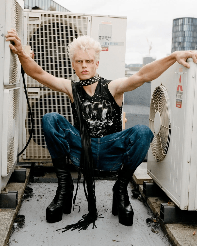 Androgynous model with blonde hair and platform boots sits atop an air conditioning unit, wearing a rock t-shirt and blue jeans.