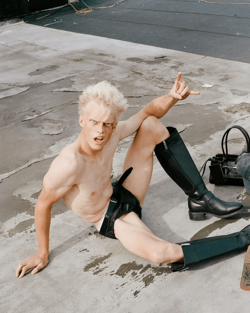 Shirtless male model with blond hair sitting on the ground in black riding boots, posing with an open mouth and hand gesture.