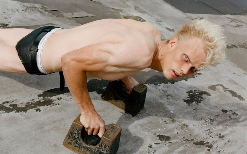 Shirtless man with blond hair and leather shorts doing push-ups on concrete using metal weights as handles, outdoors on an overcast day.