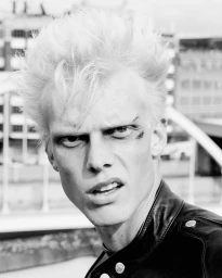 Black and white close-up portrait of a man with spiky blonde hair and a cut above his eye wearing a black leather jacket.