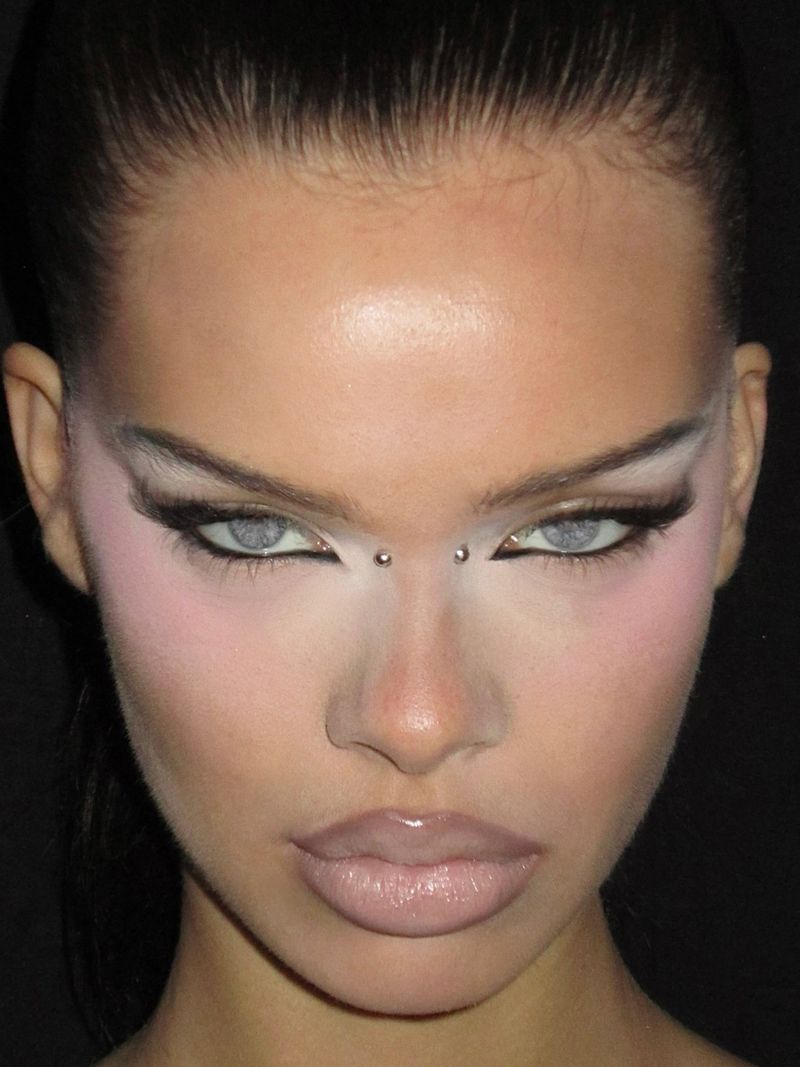 Close-up portrait of a young adult woman with creative makeup including white eyeshadow and dark eyeliner, fair skin, and a black background.