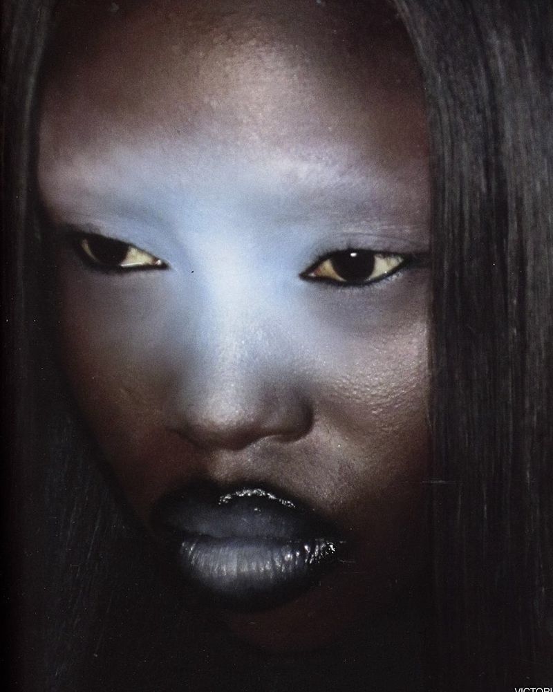 Portrait of an adult female with striking blue makeup on her forehead and chin, featuring glossy dark lipstick and sleek, straight hair.