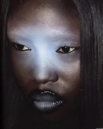 Portrait of an adult female with striking blue makeup on her forehead and chin, featuring glossy dark lipstick and sleek, straight hair.
