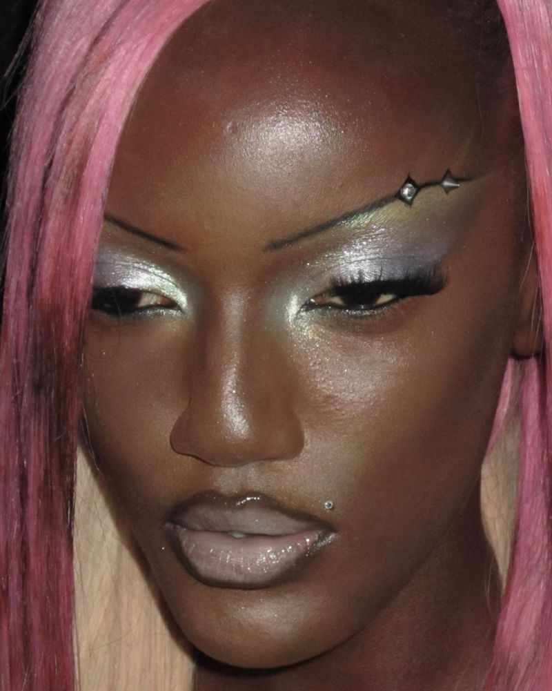 Close-up portrait of a Black adult female with pink hair, creative eye makeup, and a lip piercing, against a dark background.
