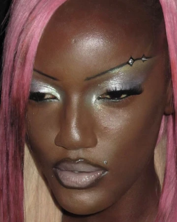 Close-up portrait of a Black adult female with pink hair, creative eye makeup, and a lip piercing, against a dark background.