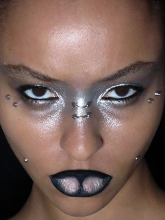 Close-up shot of an adult female with bold silver and black makeup, silver facial jewelry, and studs adorning the face against a dark backdrop.