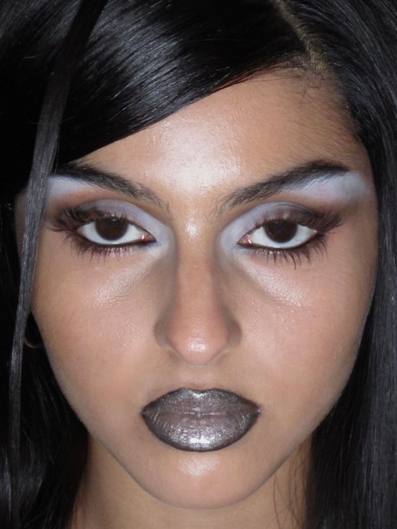 Close-up portrait of a woman with heavy makeup, including bleached eyebrows, dramatic eyeshadow, and metallic silver lipstick, looking directly at the camera.