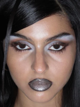 Close-up portrait of a woman with heavy makeup, including bleached eyebrows, dramatic eyeshadow, and metallic silver lipstick, looking directly at the camera.