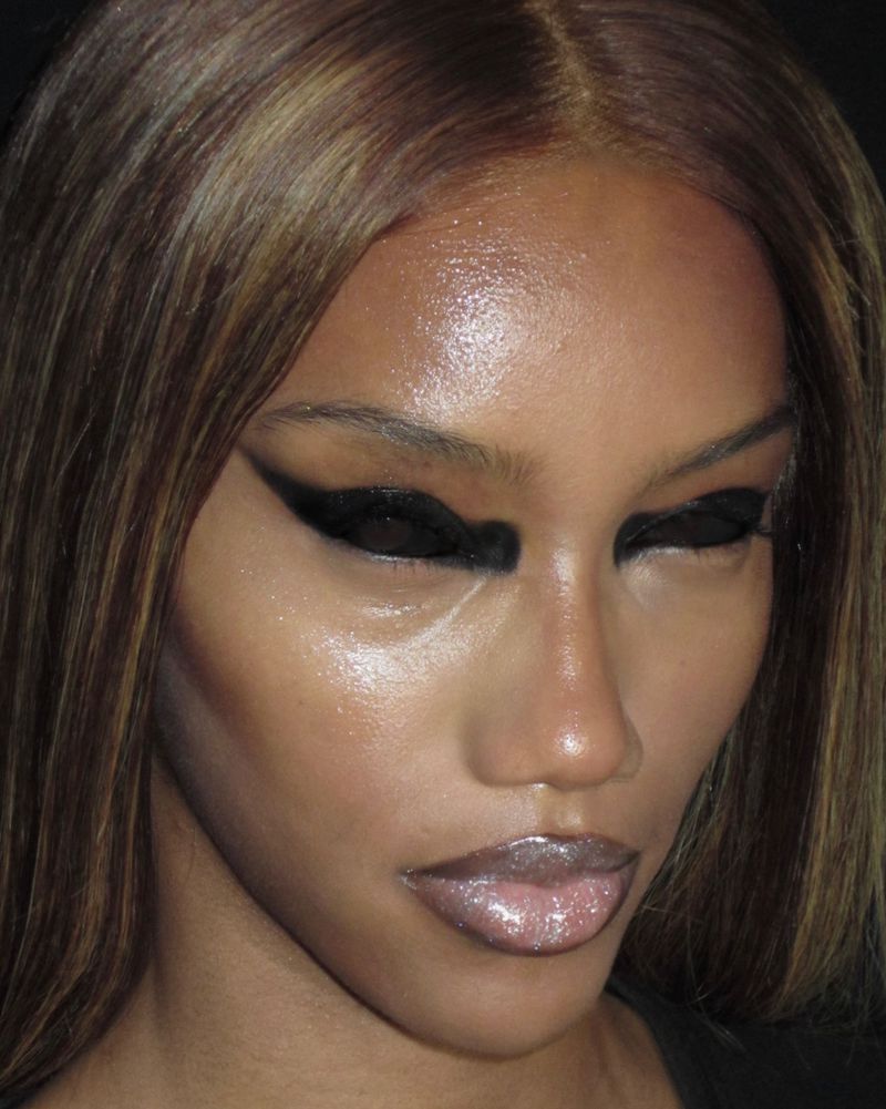 Close-up portrait of an adult female with bronze hair and exaggerated black eye makeup, highlighting her glossy lips and makeup application.
