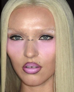 Close-up beauty portrait of a blonde model with bleached eyebrows, pink blush, face piercings, and lilac lipstick.