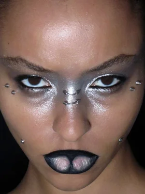 Close-up beauty portrait of a person featuring metallic silver eye makeup, dark lipstick, facial piercings, and dark background