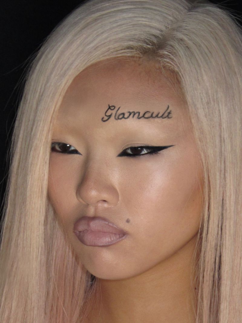 Close-up beauty portrait of an East Asian model with platinum hair, sharp winged eyeliner, and Glamcult scripted above the eyebrow.