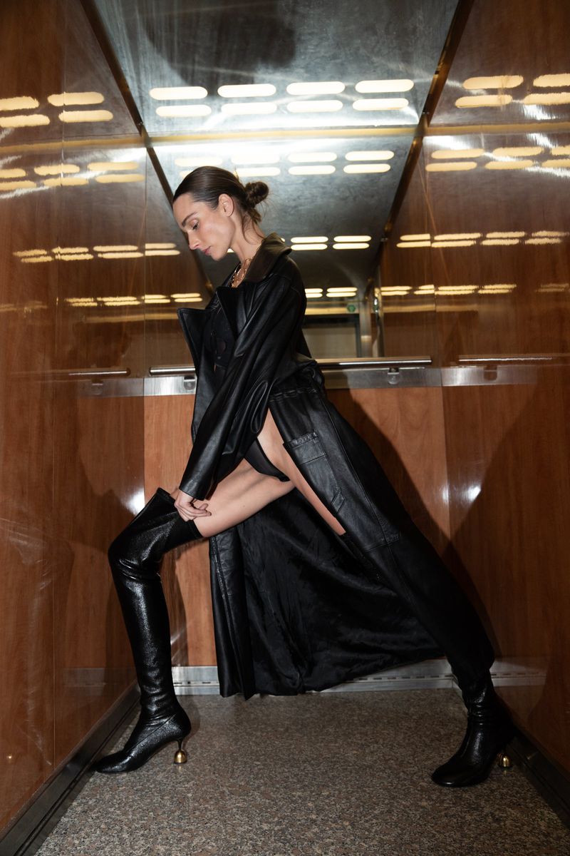A woman in a black coat and high boots poses in an elevator with wood-paneled walls and a reflective ceiling.