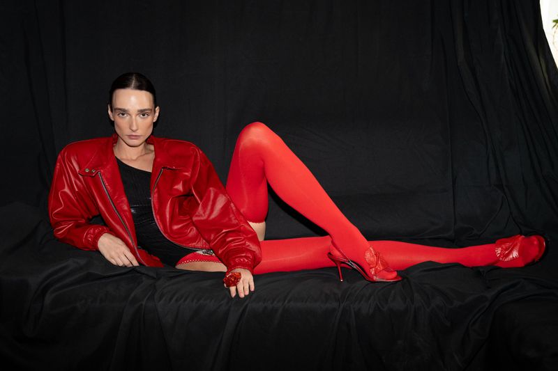A fashion photo of a model in a red jacket, stockings, and high heels, lying on a black surface, against a black backdrop.