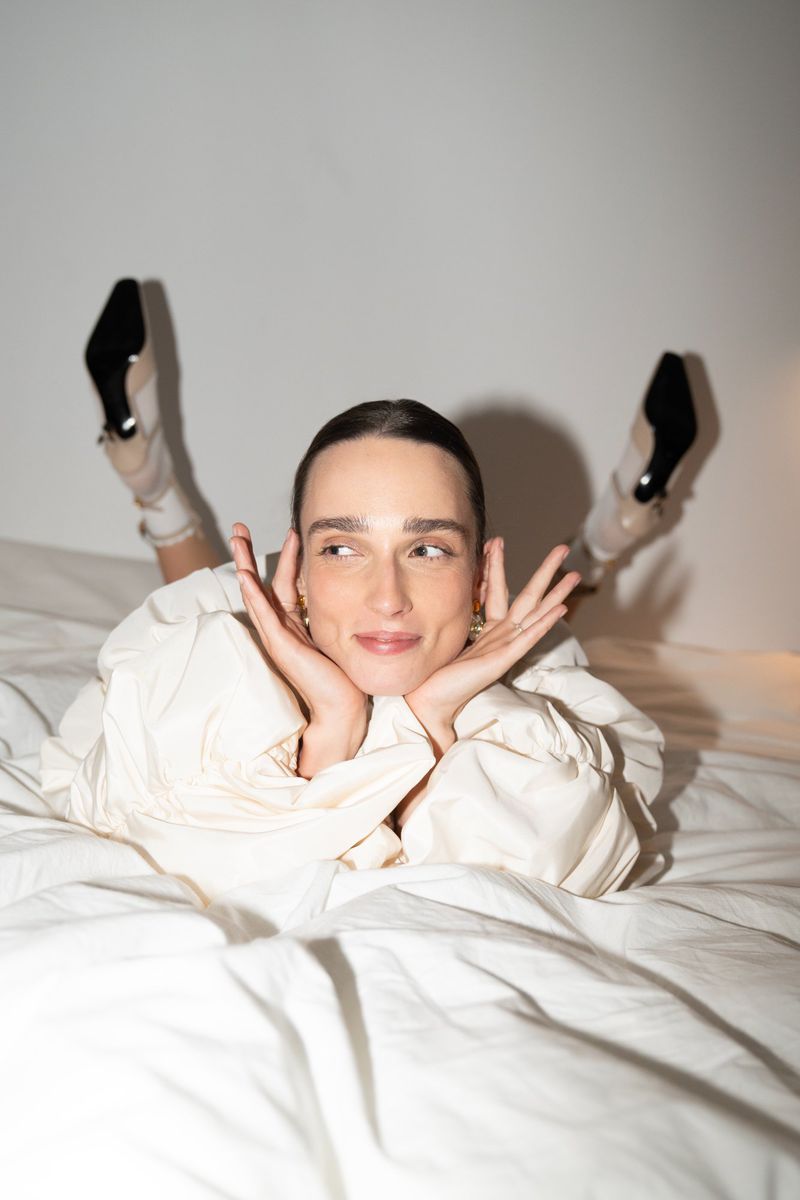 An adult woman with dark hair lies on a white bed with her head propped up, wearing a cream-colored jacket and black heels.
