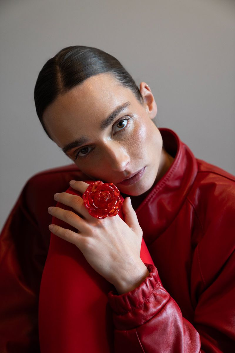 Close-up studio portrait of an adult woman in a red outfit with a large, red flower ring resting her head on her hand.