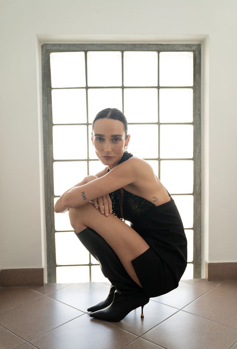 Woman in black dress and boots squats in front of window with tiled floor in neutral interior setting, looking at the camera.