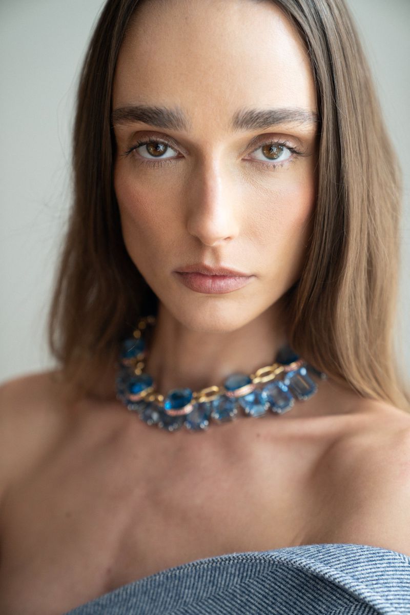 Close-up portrait of an adult woman with blue gemstone necklace, featuring soft lighting and a neutral background.