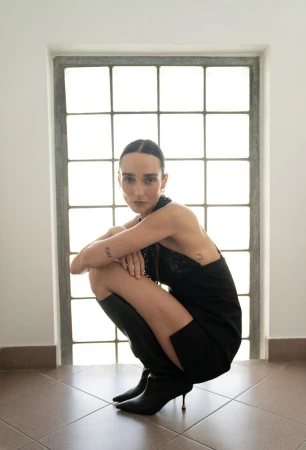 Woman wearing a black dress and boots crouching in front of a grid window for Flanelle Magazine