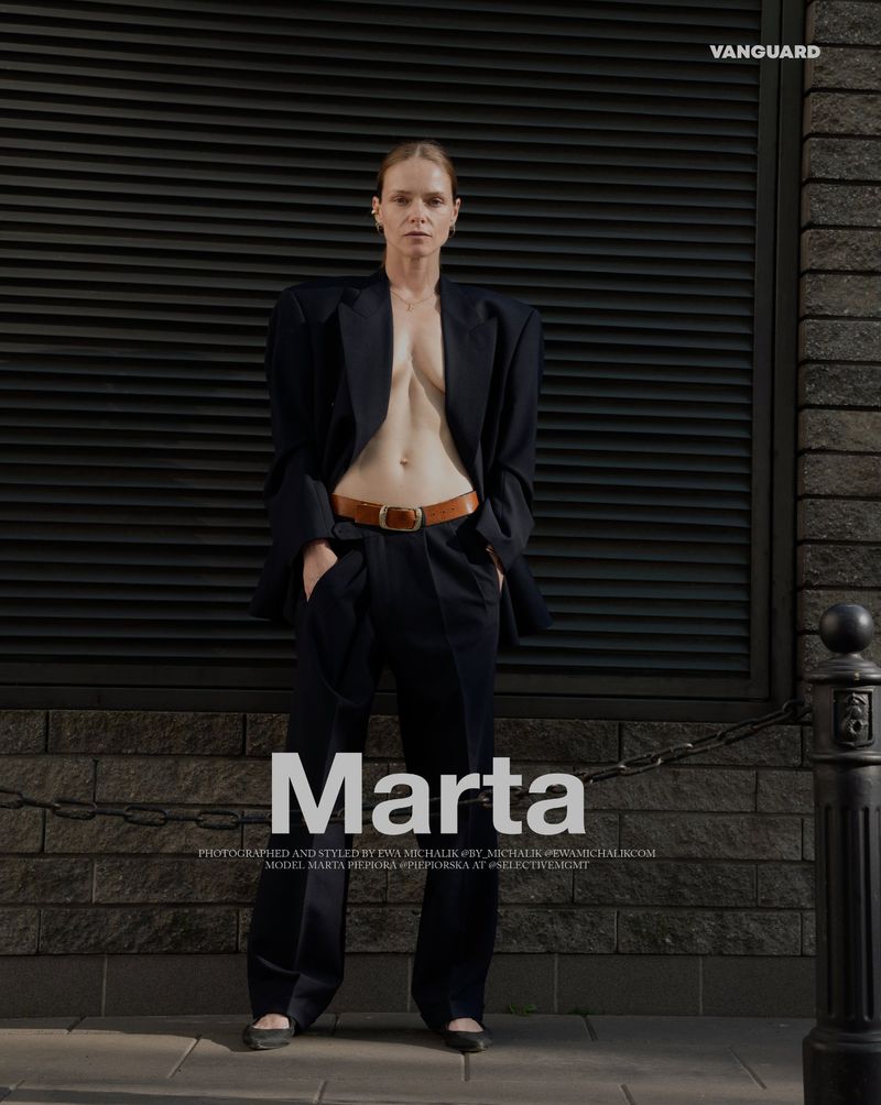 Fashion editorial shot: a slim adult woman poses in an oversized navy suit with a brown belt and black shoes against a dark brick wall.