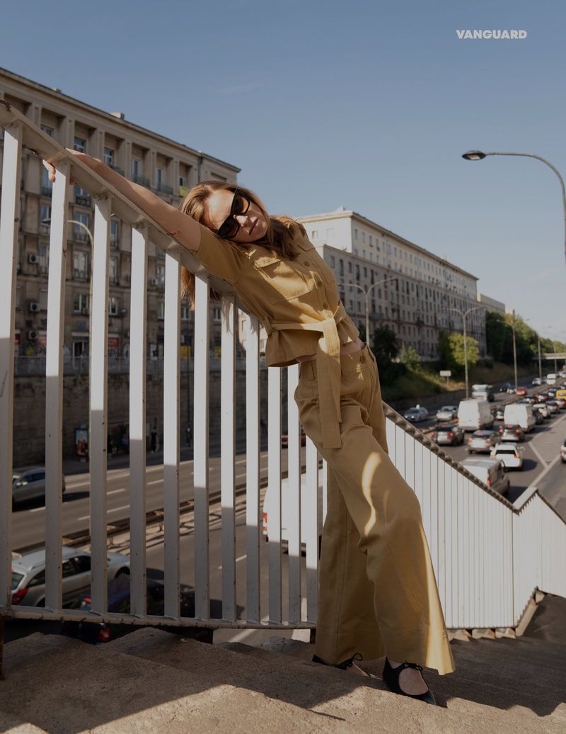 Fashion editorial shot: a stylish adult woman leans against a railing in wide-leg pants and a matching top in an urban setting with city traffic.