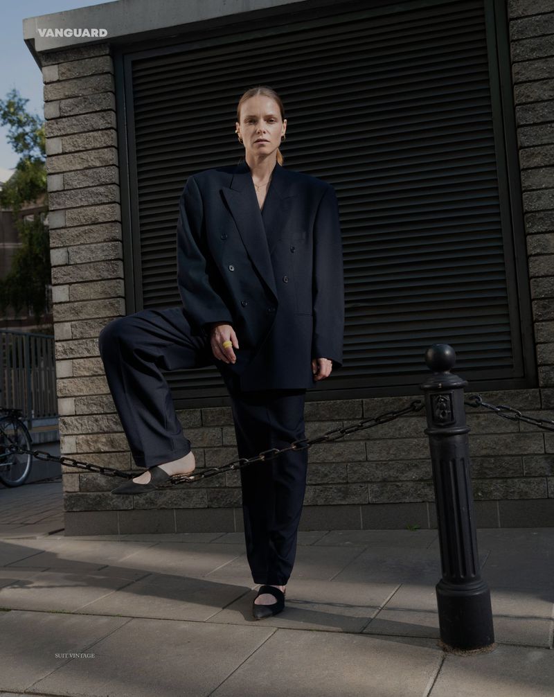 Fashionable woman in an oversized navy suit strikes a pose on a city street with textured brick buildings and metal bollards with chain.