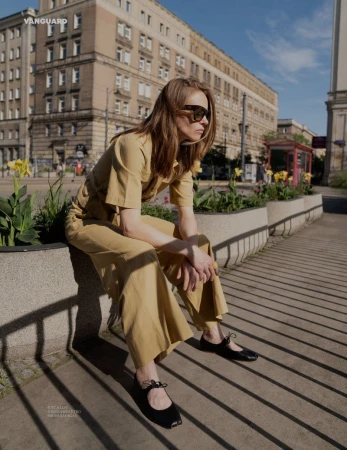 Woman in tan jumpsuit and sunglasses sits on a wall in a city with a vintage building in the background on a sunny day.