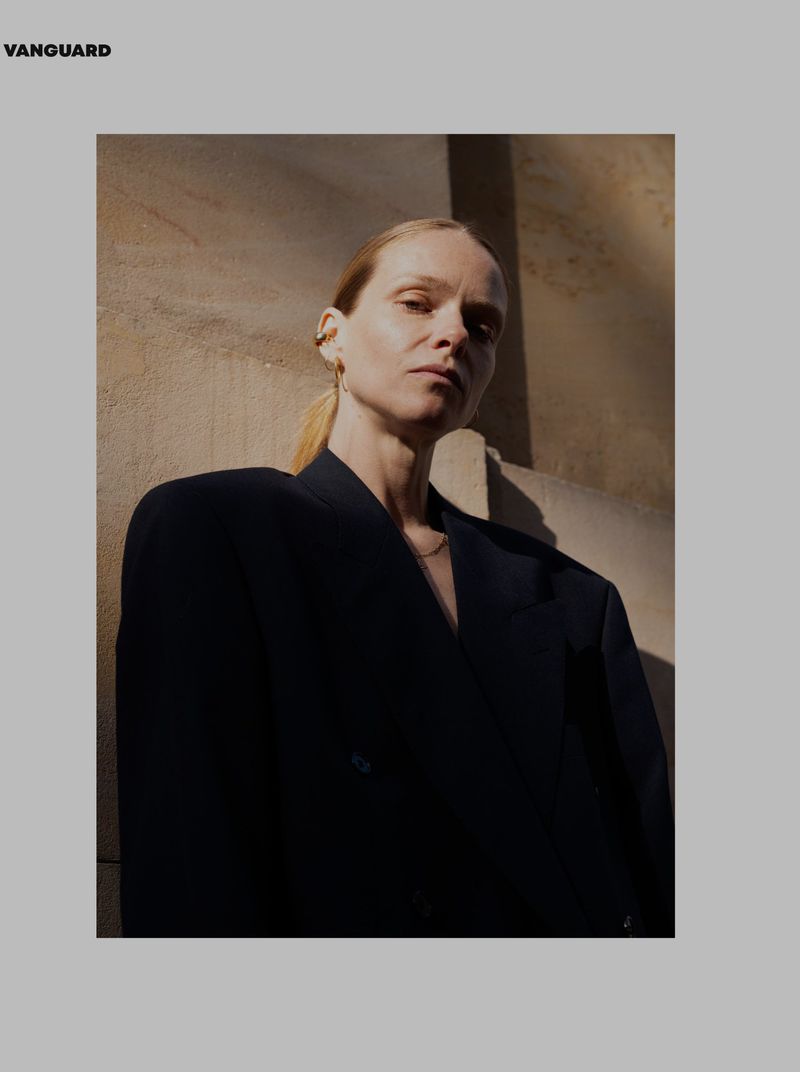 Blonde woman with ponytail wears a dark blazer in an eye-level portrait against a textured wall with natural lighting.
