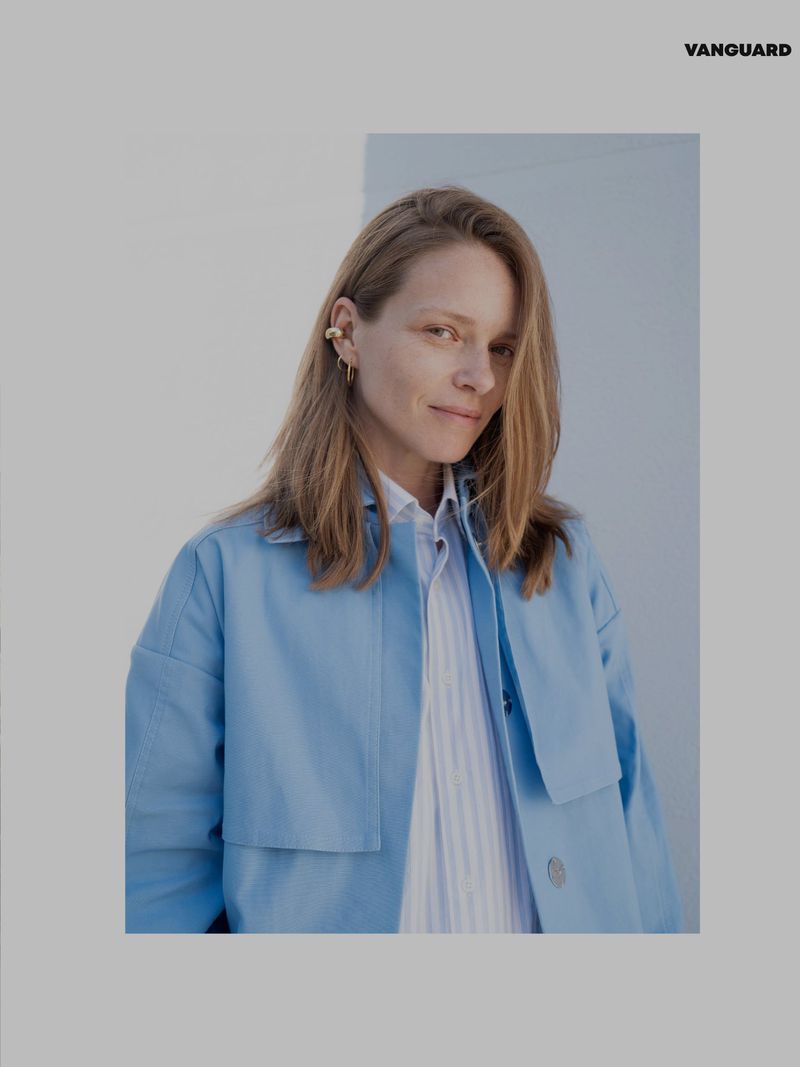 Portrait of a blond adult woman in a light blue jacket and striped shirt against a split white and light blue background in a photo studio.