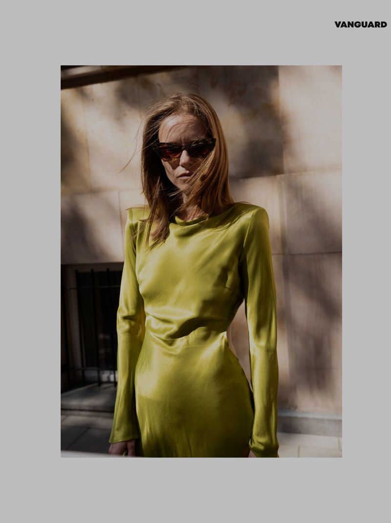 A fair-skinned adult female model poses in a chartreuse, long-sleeved dress in front of a textured wall in an outdoor setting.