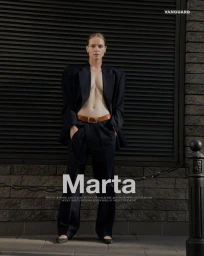 Fashion model standing outdoors wearing an open oversized black suit jacket with matching trousers and a brown leather belt.