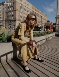 A woman wearing dark sunglasses, a tan short-sleeved jumpsuit, and black flats sits on a concrete planter along a city street.