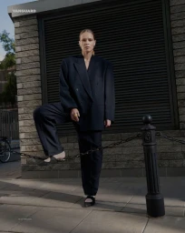 Model wearing an oversized dark suit posing outdoors with one foot resting on a chain fence post for Vanguard Magazine project Marta.